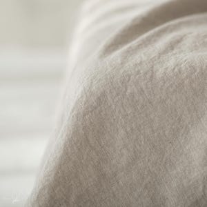 May include: A close-up view of a light beige fabric with a visible woven texture. The material features a natural, slightly wrinkled appearance with soft folds cascading down, highlighting the organic weave and neutral color of the textile.