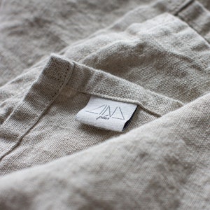 May include: A beige, textured fabric with a visible weave, featuring a white rectangular tag sewn onto the corner. The tag displays a black graphic of a triangle with the text ZINA plus printed underneath in a simple, clean, black font.