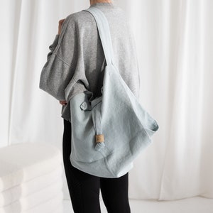 May include: A light blue linen tote bag with a long strap. The bag is worn over the shoulder of a person wearing a grey sweatshirt and black leggings.