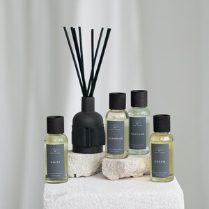 Puede incluir: Cinco botellas de vidrio de aceite difusor con tapas y etiquetas negras. Las botellas están dispuestas sobre una superficie blanca texturizada. Las etiquetas dicen "Unity", "Glamour", "Fortune" y "Dream". Una botella difusora negra con cañas negras está en el fondo.