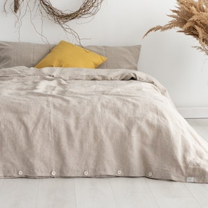 Puede incluir: Una funda n&oacute;rdica de lino beige con cierre de botones. La funda n&oacute;rdica est&aacute; en una cama con una almohada amarilla y dos fundas de almohada de lino beige. La cama est&aacute; en una habitaci&oacute;n con una pared blanca y una corona de ramas secas colgada sobre la cama.
