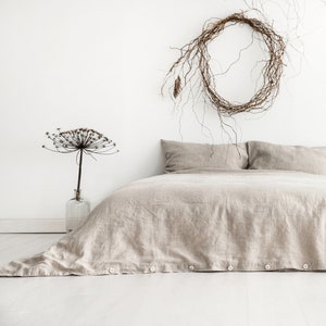 Puede incluir: Una funda n&oacute;rdica de lino beige con cierres de bot&oacute;n en una cama con dos almohadas de lino beige. Un arreglo de flores secas est&aacute; sobre una mesa a la izquierda de la cama. Una corona de ramas cuelga en la pared detr&aacute;s de la cama.