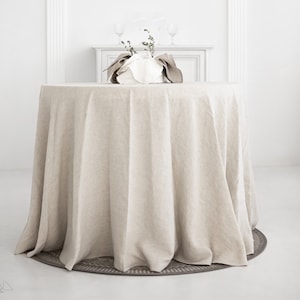 May include: A round, beige linen tablecloth draped over a table in a white room. The tablecloth has a natural, slightly wrinkled texture and hangs down to the floor.