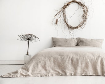 May include: A beige linen duvet cover with button closures on a bed with two beige linen pillows. A dried flower arrangement is on a table to the left of the bed. A twig wreath hangs on the wall behind the bed.