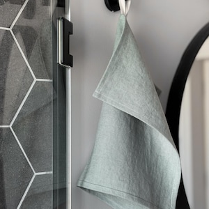 May include: A sage green hand towel with a visible woven texture, featuring a white fabric hanging loop. The towel is suspended from a matte black circular wall hook, showcasing a simple rectangular shape with hemmed edges and a soft, muted color.