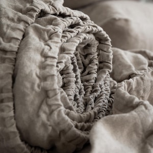 May include: A beige fitted sheet made of textured fabric, rolled up to reveal the elasticized edge. The material has a visible weave and a soft, matte appearance. The sheet is gathered in tight, uniform folds along the circular elastic border.