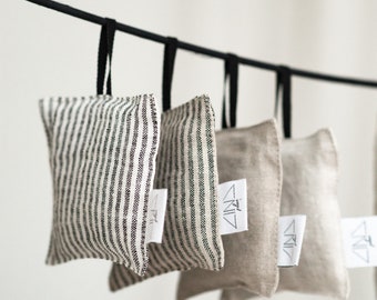 May include: Five fabric sachets hanging from a black metal rod. The sachets are made of linen and have a striped pattern. Each sachet has a white label with the word "Zinz" printed on it.