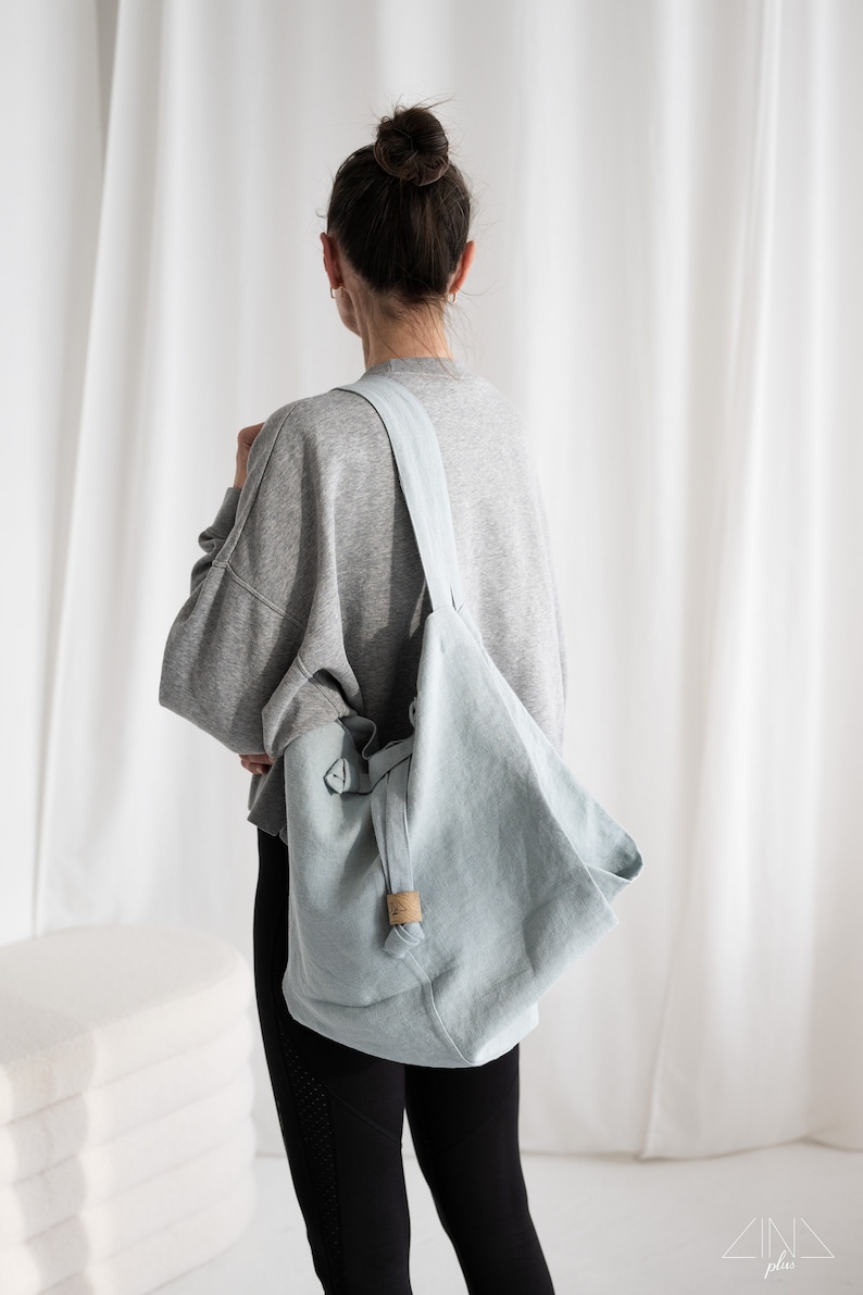 May include: A light blue linen tote bag with a long strap. The bag is worn over the shoulder of a person wearing a grey sweatshirt and black leggings.