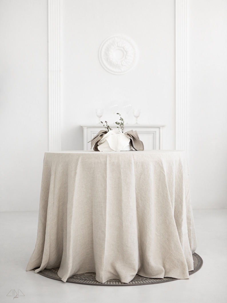 May include: A round, beige linen tablecloth draped over a table in a white room. The tablecloth has a natural, slightly wrinkled texture and hangs down to the floor.
