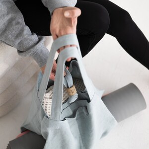 May include: A light blue tote bag with a white mesh bag inside. The bag has a brown leather tag with the word "plus" on it. The bag is sitting on a gray yoga mat.