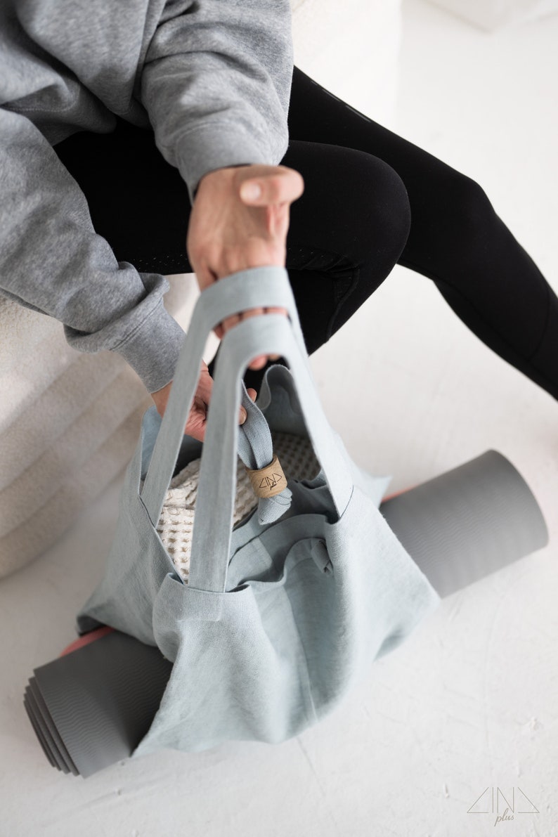 May include: A light blue tote bag with a white mesh bag inside. The bag has a brown leather tag with the word "plus" on it. The bag is sitting on a gray yoga mat.