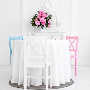 Puede incluir: Una mesa blanca con un mantel blanco y un centro de mesa floral rosa. Hay tres sillas alrededor de la mesa, una azul, una rosa y una blanca.