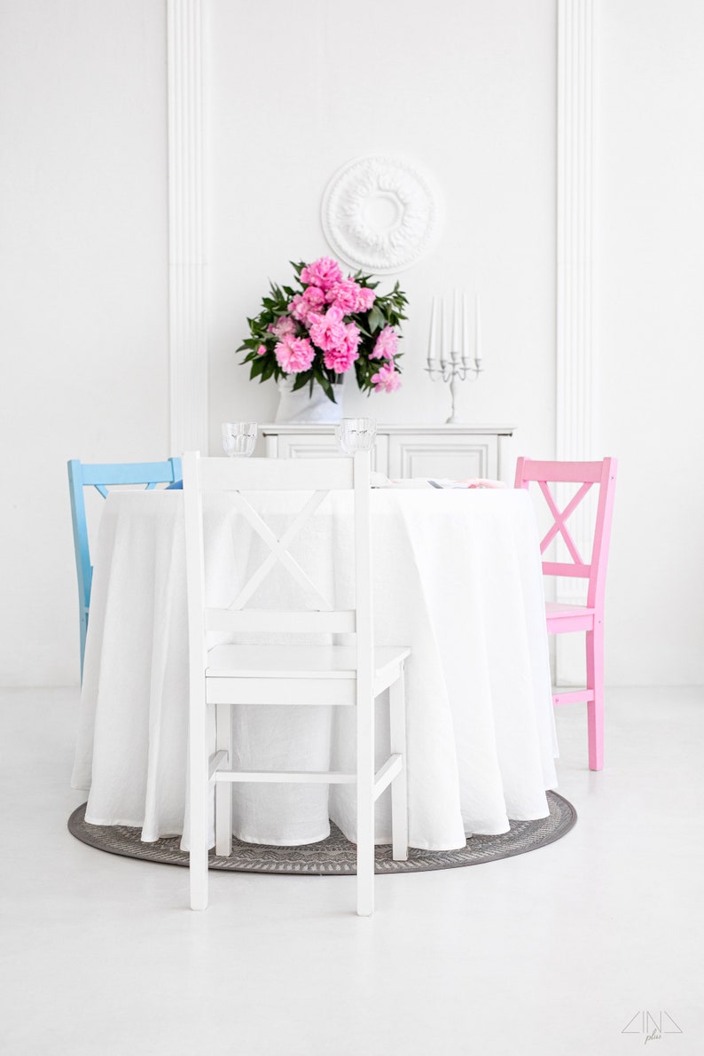 Puede incluir: Una mesa blanca con un mantel blanco y un centro de mesa floral rosa. Hay tres sillas alrededor de la mesa, una azul, una rosa y una blanca.