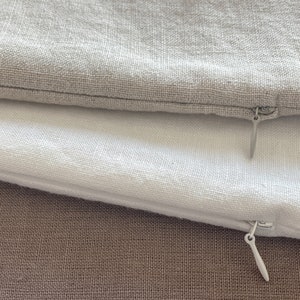 May include: Two pillowcases with zippers, one is white and the other is a light gray. The pillowcases are laying on a brown fabric surface.