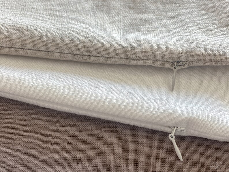May include: Two pillowcases with zippers, one is white and the other is a light gray. The pillowcases are laying on a brown fabric surface.