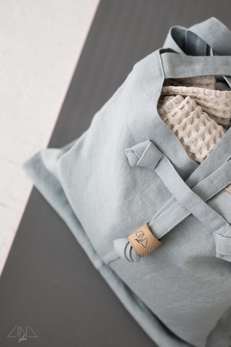 May include: A light blue linen tote bag with a wooden tag that says "CIND plus". The bag has a criss-cross strap design and is partially filled with a white mesh bag.
