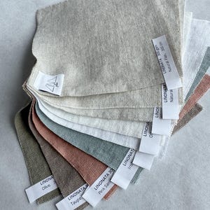 Puede incluir: Una colecci&oacute;n en abanico de muestras de tela de lino cuadradas en varios colores, incluyendo natural, blanco, oliva, topo, arena rosada e iris. Cada muestra tiene una etiqueta blanca con texto como LINONATA 300 gsm, Natural Flax, 300 cm.