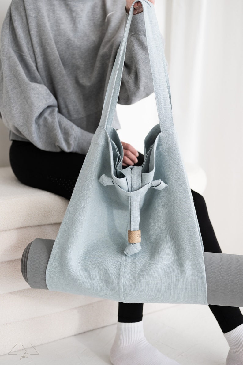 May include: A light blue linen yoga mat bag with a drawstring closure and a wooden toggle. The bag is being held by a person wearing black leggings and white socks.