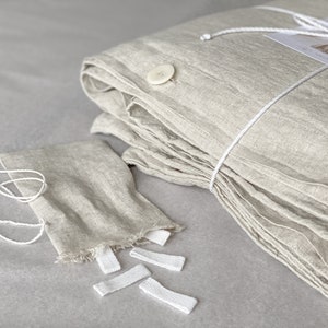Puede incluir: Una funda n&oacute;rdica de lino doblada en color beige claro con lazos y botones blancos. La funda n&oacute;rdica est&aacute; doblada y atada con una cuerda blanca. Tambi&eacute;n hay varios lazos blancos sobre la mesa.