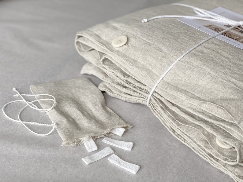 Puede incluir: Una funda n&oacute;rdica de lino doblada en color beige claro con lazos y botones blancos. La funda n&oacute;rdica est&aacute; doblada y atada con una cuerda blanca. Tambi&eacute;n hay varios lazos blancos sobre la mesa.