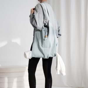 May include: A person wearing black leggings and a gray sweatshirt carries a light blue tote bag with a wooden handle. The bag contains a rolled-up yoga mat and a white towel. The person is standing on a gray yoga mat with a pink edge.