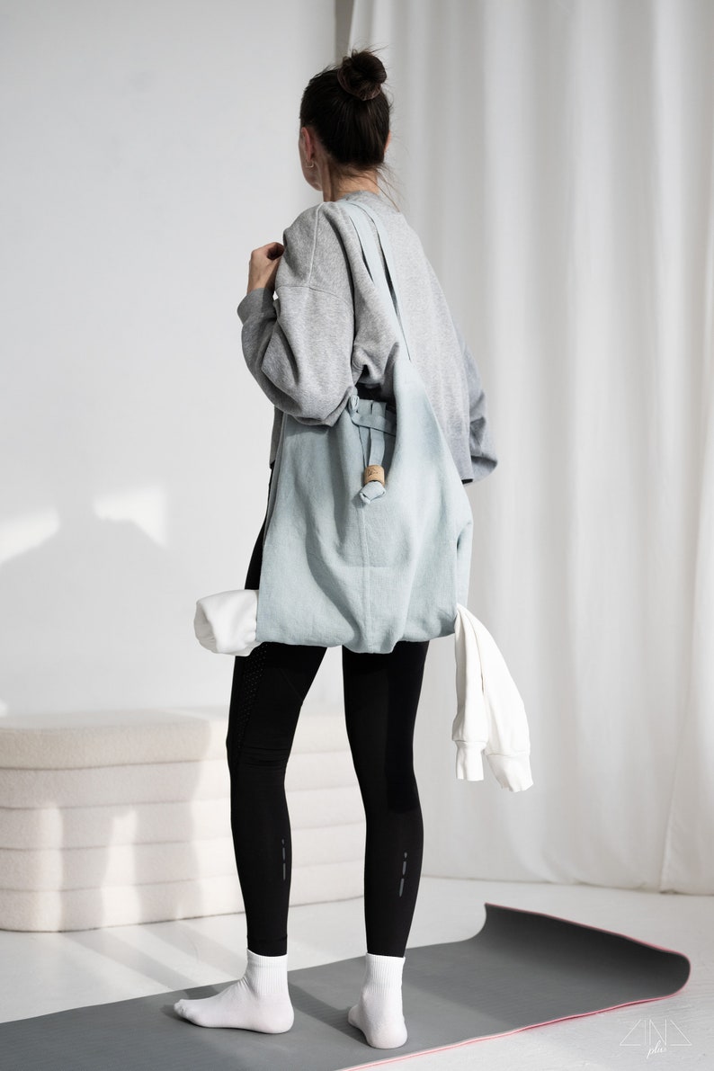 May include: A person wearing black leggings and a gray sweatshirt carries a light blue tote bag with a wooden handle. The bag contains a rolled-up yoga mat and a white towel. The person is standing on a gray yoga mat with a pink edge.