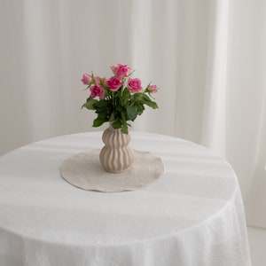 Puede incluir: Un ramo de rosas rosas en un jarr&oacute;n de cer&aacute;mica marr&oacute;n claro texturizado sobre un mantel blanco. El jarr&oacute;n tiene una forma redonda y bulbosa con nervaduras verticales.