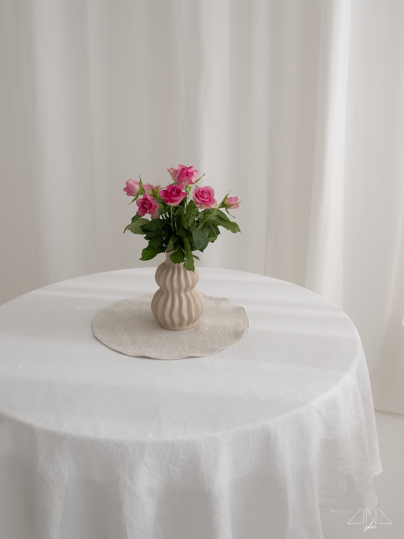 Puede incluir: Un ramo de rosas rosas en un jarr&oacute;n de cer&aacute;mica marr&oacute;n claro texturizado sobre un mantel blanco. El jarr&oacute;n tiene una forma redonda y bulbosa con nervaduras verticales.