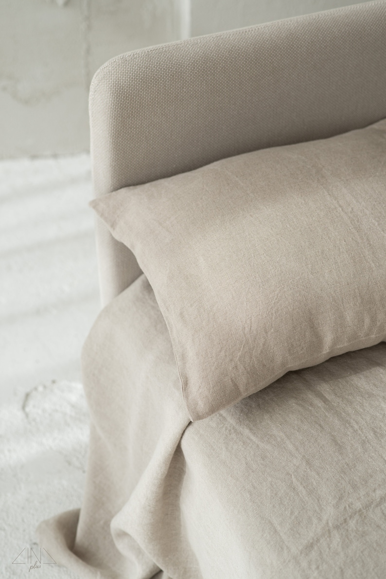 May include: A close-up of a beige linen bed with a pillow. The bed is made with a soft, natural fabric and has a simple, elegant design.