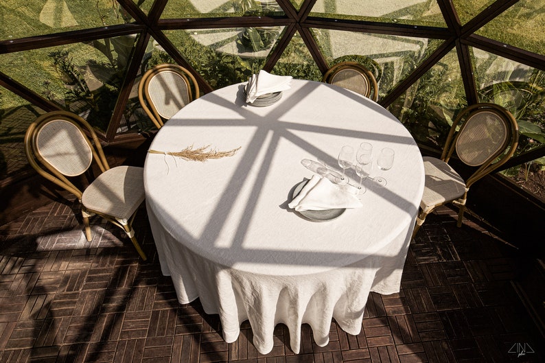 May include: A white tablecloth covers a round table set for four with white napkins, clear glasses, and a single dried grass stalk. The table is in a geodesic dome with a wooden floor.