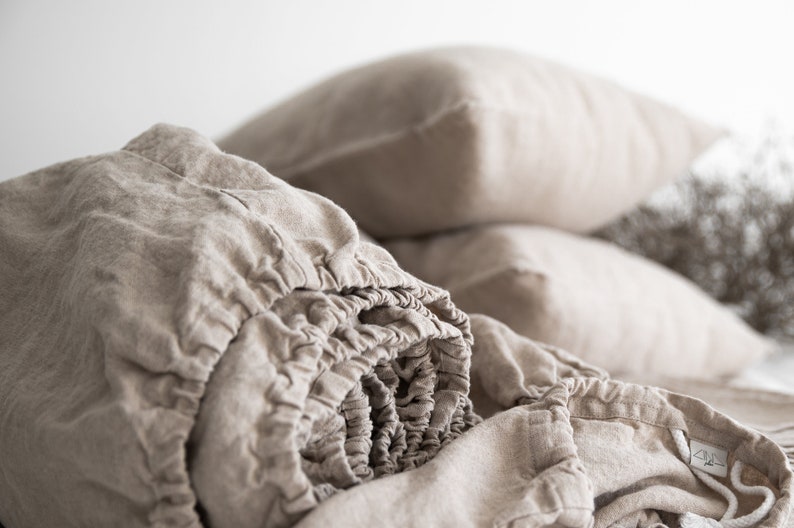 May include: A close-up of a beige linen fitted sheet, rolled up and ready to be placed on a bed. The sheet is made of natural linen fabric, known for its breathability and durability.