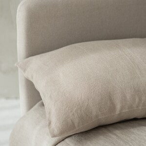 May include: A close-up of a beige linen pillow resting on a beige linen bed. The pillow is slightly wrinkled and has a textured surface.