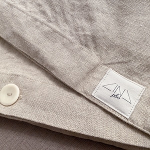 Op de afbeelding: Een close-up van een beige linnen dekbedovertrek met een witte knoop en een wit label met de tekst "ZINA plus" in zwarte letters.