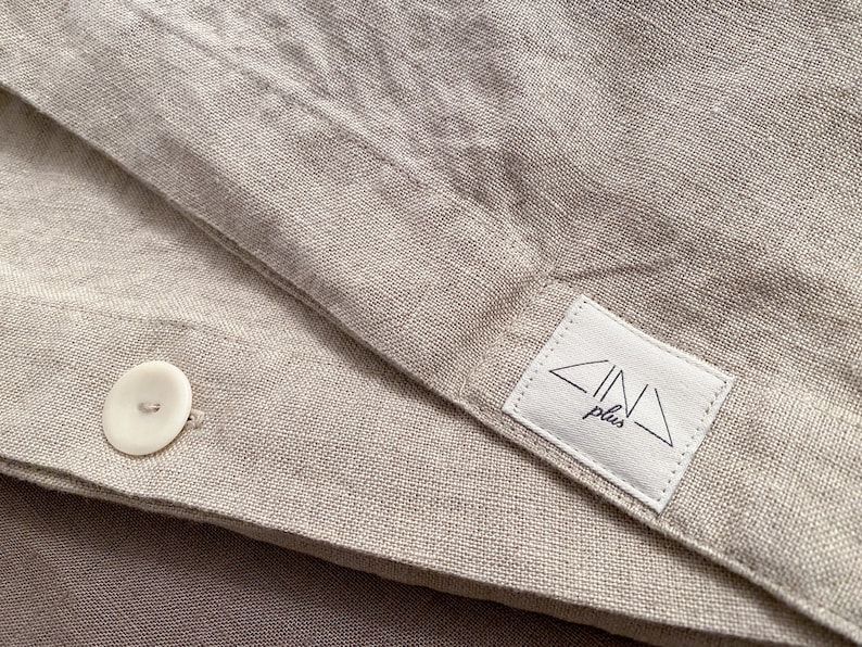 Op de afbeelding: Een close-up van een beige linnen dekbedovertrek met een witte knoop en een wit label met de tekst "ZINA plus" in zwarte letters.