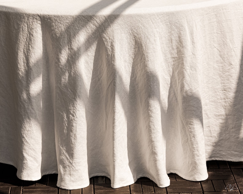 May include: A white linen tablecloth with a textured surface. The tablecloth is draped over a round table and has soft folds and creases. The tablecloth is illuminated by sunlight, creating shadows and highlights.