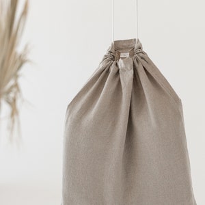 Puede incluir: Una bolsa de cord&oacute;n de lino beige con cordones blancos que cuelgan de la parte superior. La bolsa est&aacute; vac&iacute;a y cuelga sobre un fondo blanco.