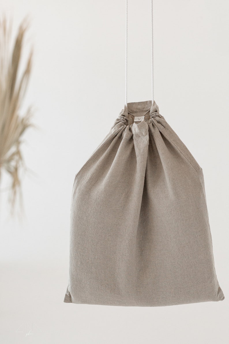 Puede incluir: Una bolsa de cord&oacute;n de lino beige con cordones blancos que cuelgan de la parte superior. La bolsa est&aacute; vac&iacute;a y cuelga sobre un fondo blanco.