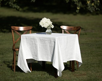May include: A white linen tablecloth covers a square wooden table with two wooden chairs. A small glass vase with white flowers sits in the center of the table.