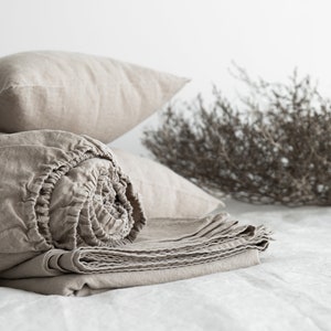 May include: A rolled up beige linen fitted sheet with a beige linen pillow on top of it. The sheet is folded neatly and the pillow is slightly wrinkled. The background is a white wall with a dried branch in the background.