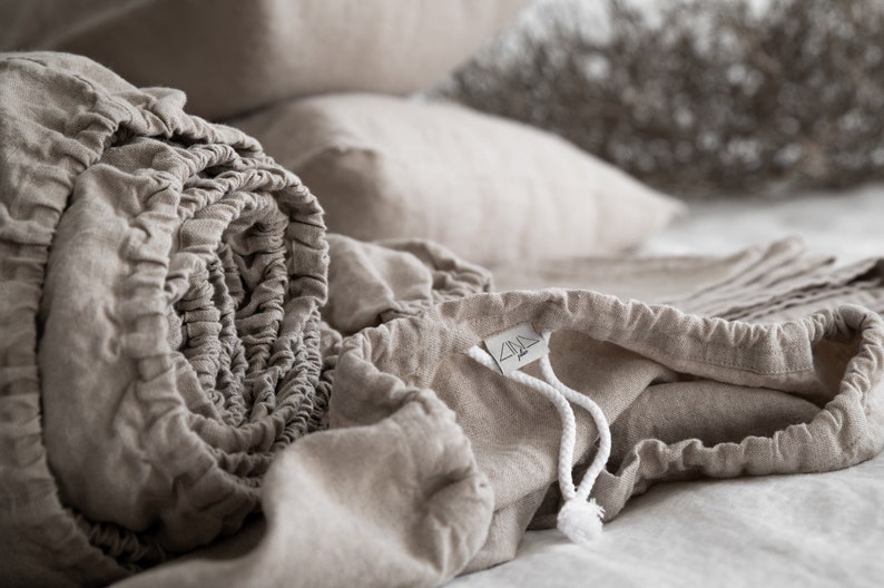 May include: A close-up of a beige linen fitted sheet with a drawstring bag. The sheet is rolled up and the bag is partially open, revealing a white drawstring.