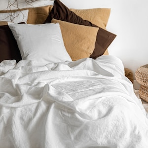 K&ouml;nnte beinhalten: Ein Bett mit wei&szlig;er Leinenbettw&auml;sche, einem wei&szlig;en Kissen und mehreren Kissen in Braun- und Beiget&ouml;nen. &Uuml;ber dem Bett h&auml;ngt ein dekorativer Zweigkranz. Die Szene vermittelt ein Gef&uuml;hl von Ruhe und Komfort.