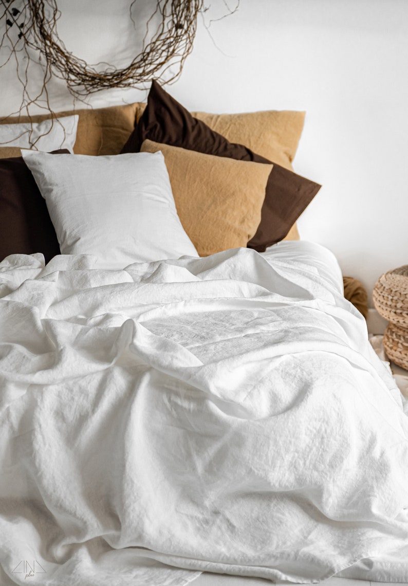 K&ouml;nnte beinhalten: Ein Bett mit wei&szlig;er Leinenbettw&auml;sche, einem wei&szlig;en Kissen und mehreren Kissen in Braun- und Beiget&ouml;nen. &Uuml;ber dem Bett h&auml;ngt ein dekorativer Zweigkranz. Die Szene vermittelt ein Gef&uuml;hl von Ruhe und Komfort.