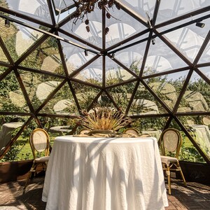 May include: A white tablecloth covers a round table set for a meal in a glass geodesic dome. Two chairs are placed on either side of the table. The dome is made of clear glass panels and has a metal frame. The dome is surrounded by green foliage.