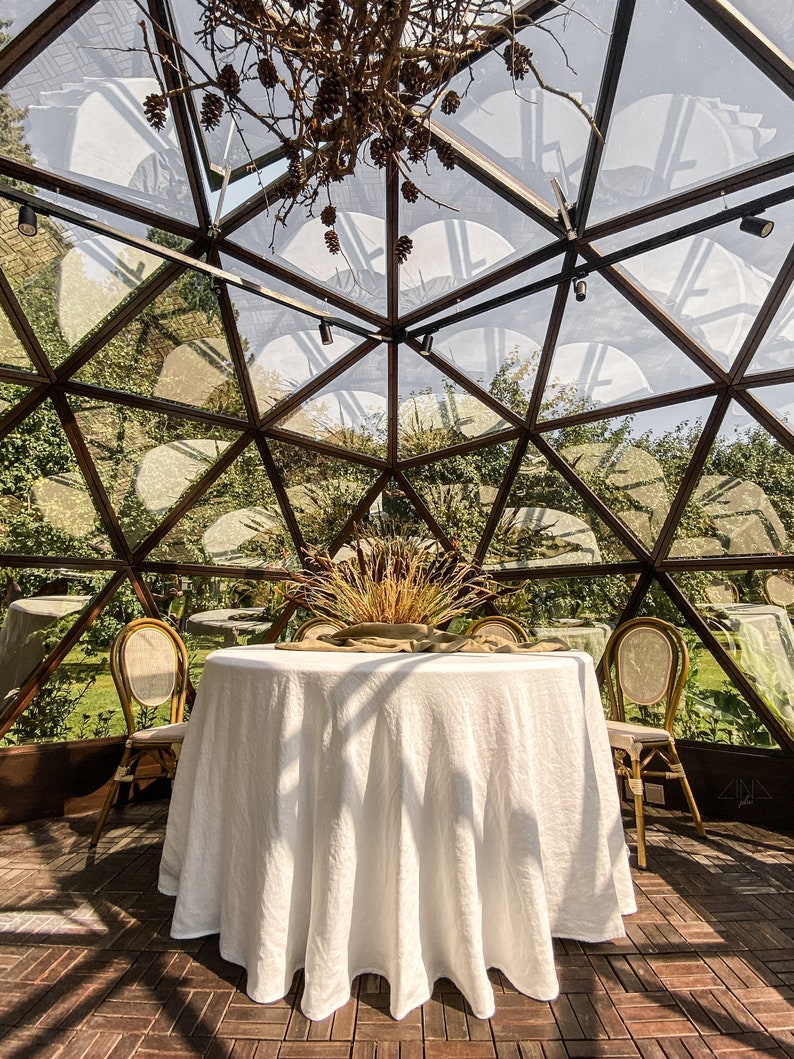 May include: A white tablecloth covers a round table set for a meal in a glass geodesic dome. Two chairs are placed on either side of the table. The dome is made of clear glass panels and has a metal frame. The dome is surrounded by green foliage.