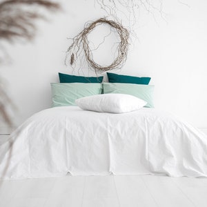 May include: A bed with white linens, teal and mint green pillows, and a white pillow. A decorative wreath hangs above the bed. The room has white walls and flooring. The image has a minimalist, modern aesthetic.