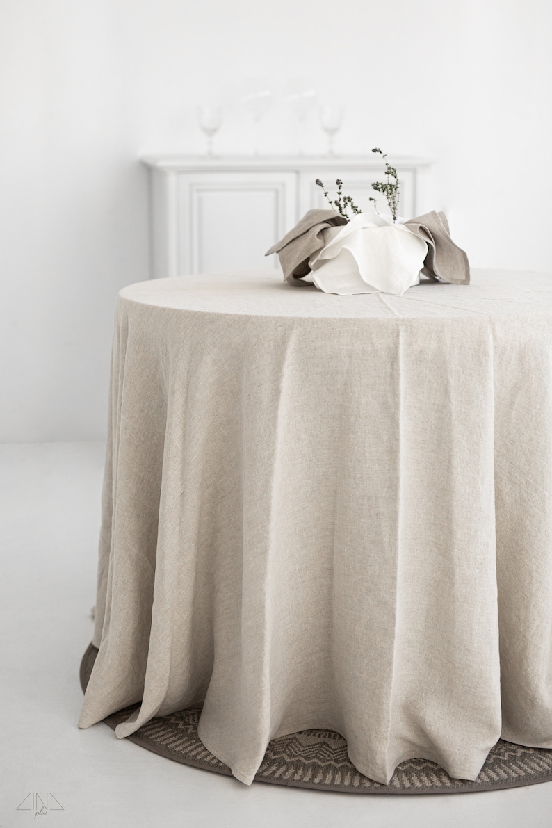 May include: A round, beige linen tablecloth draped over a table with a white ceramic bowl filled with dried greenery in the center.