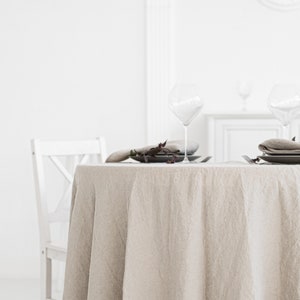 Puede incluir: Un mantel redondo de lino beige drapeado sobre una mesa blanca con dos cubiertos. Cada cubierto incluye un plato blanco, una copa de vino de vidrio y una peque&ntilde;a rama de vegetaci&oacute;n.