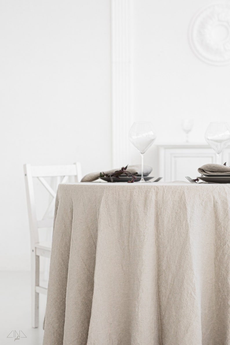 Puede incluir: Un mantel redondo de lino beige drapeado sobre una mesa blanca con dos cubiertos. Cada cubierto incluye un plato blanco, una copa de vino de vidrio y una peque&ntilde;a rama de vegetaci&oacute;n.