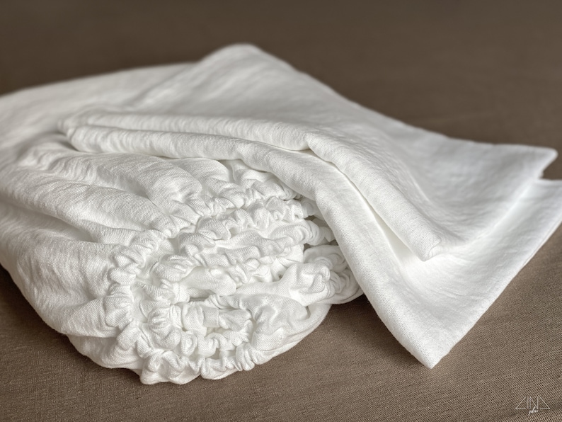 May include: A white linen fitted sheet with elastic all around the edges. The sheet is folded and appears soft and comfortable.