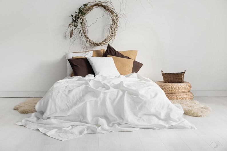 K&ouml;nnte beinhalten: Ein Bett mit wei&szlig;er Leinenbettw&auml;sche, akzentuiert durch braune, beige und wei&szlig;e Kissen. Ein gewebter Kranz h&auml;ngt &uuml;ber dem Bett. Ein kleiner Weidenkorb steht auf zwei gewebten Ottomanen.